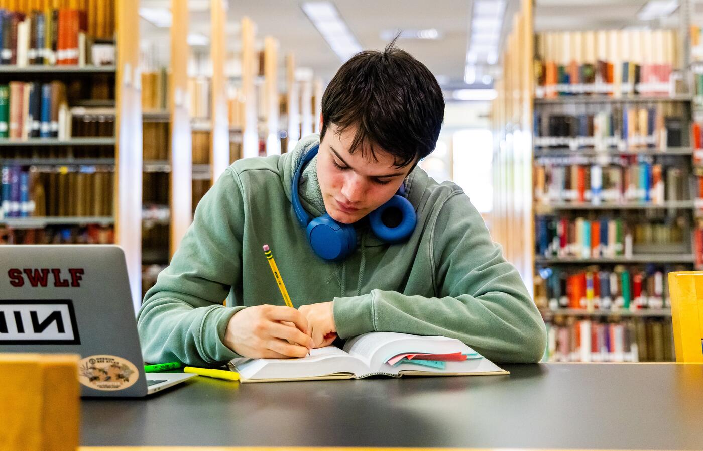 A student sitting at a table in the library writing in a textbook with their laptop open.