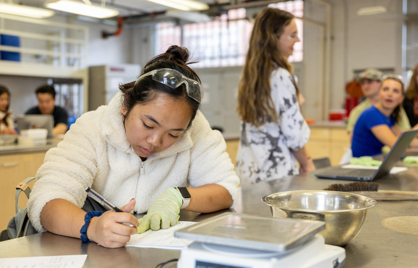 Student writing in a Lab. Laptop and other students in the background.