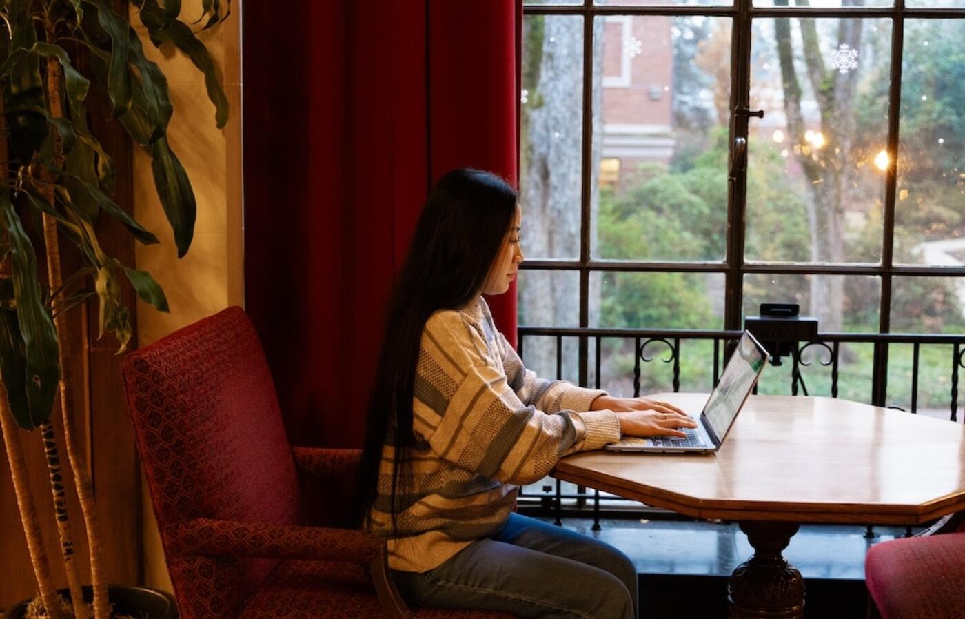 Student sitting at a table in the Memorial Union in front of a window. She has her laptop open and is typing.